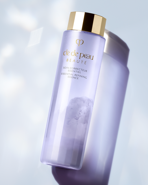 Photo of "ESSENTIAL REFINING ESSENCE" placed diagonally against a bright background, featuring a gold cap and a clear bottle in a soft purple hue.
