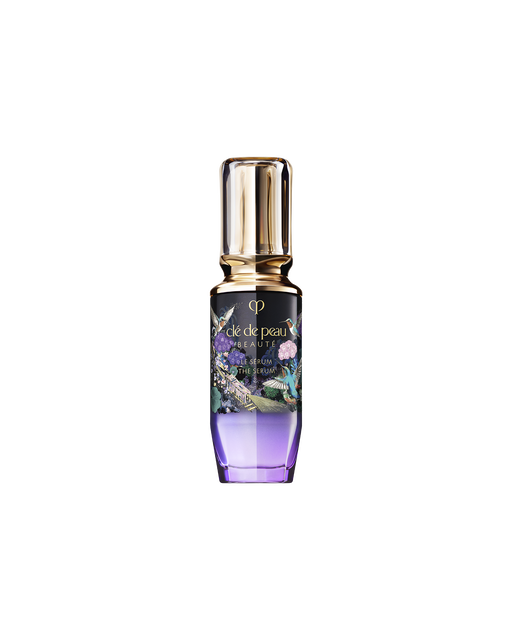Photo of the limited-edition THE SERUM bottle. The design features a gold cap and a gradient bottle from deep violet to clear. The label showcases a gold train, blue birds, and colorful flowers. SIZE: 50ml