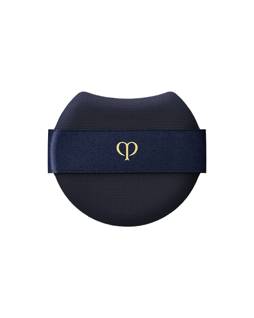 Photo of a "PUFF (RADIANT CUSHION FOUNDATION)" featuring a rounded blue-black makeup puff with a wide, dark blue ribbon across the front and the brand-logo printed in gold at the center.