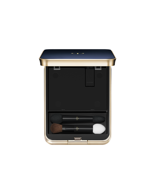 Photo of an exclusive case for "EYE COLOR QUAD" with the lid open. The blue-black lid features a gold trim. Inside, the top half has an empty compartment, while the bottom section holds two makeup applicators: a small brush and sponge-tip applicators.
