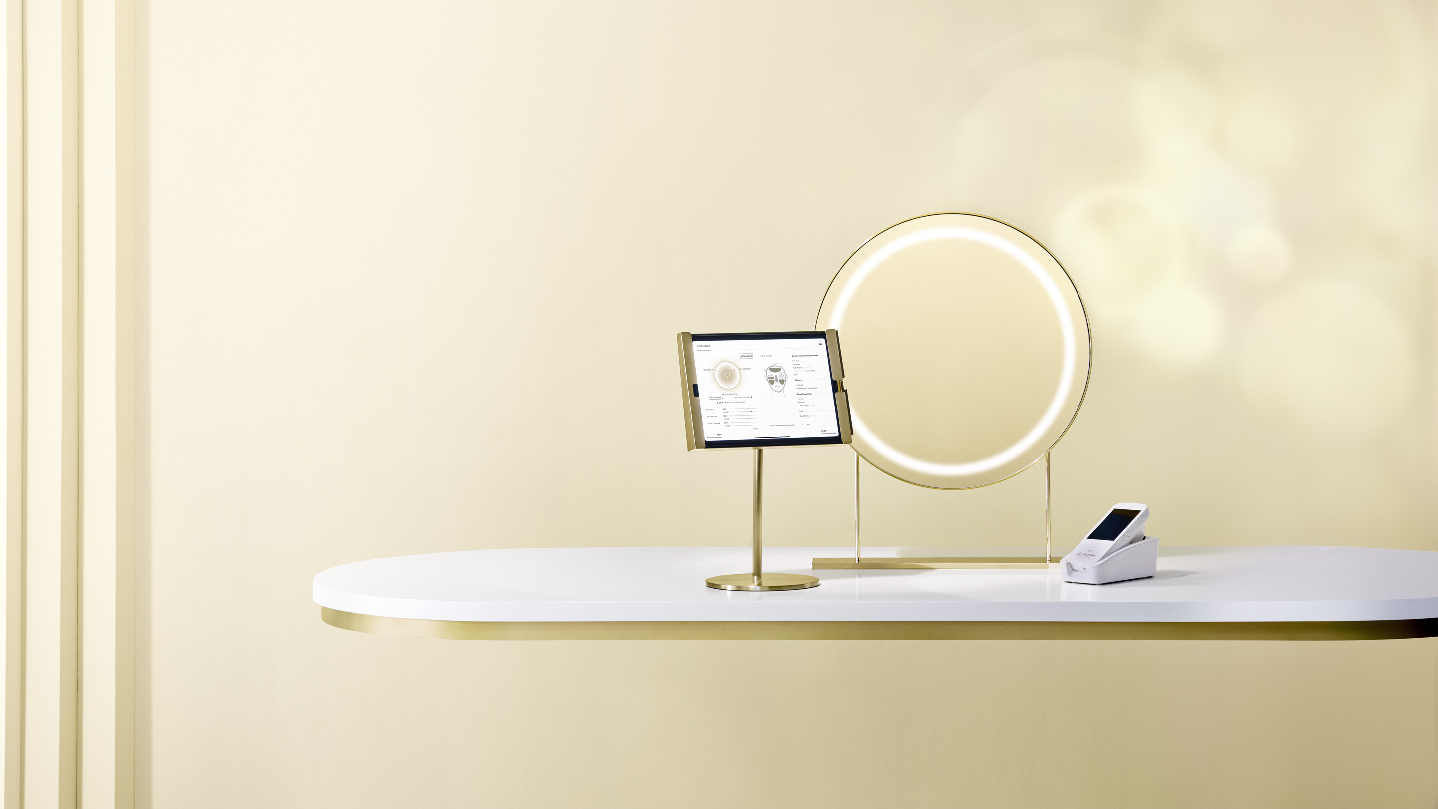 Photo of the Clé de Peau Beauté Smart Radiance Mirror setup, showing a gold-framed ring mirror, an iPad connected to a Handy Skin Sensor (HSS), and a white HSS device placed on a clean display table.