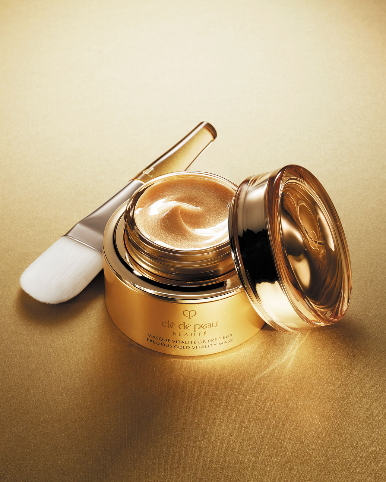 Photo of an open jar of Clé de Peau Beauté's Precious Gold Vitality Mask, showcasing its rich, creamy golden texture, with a soft, white-bristled application brush resting beside it to suggest a luxurious and pampering skincare ritual.