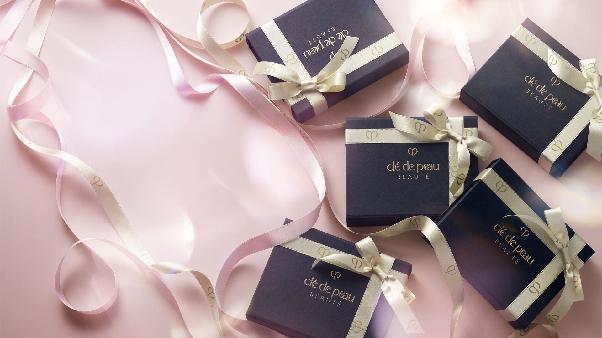 Wallpaper of Clé de Peau Beauté dark blue gift boxes arranged on a soft pink background, with champagne gold and pink ribbons featuring the brand logo elegantly intertwined.