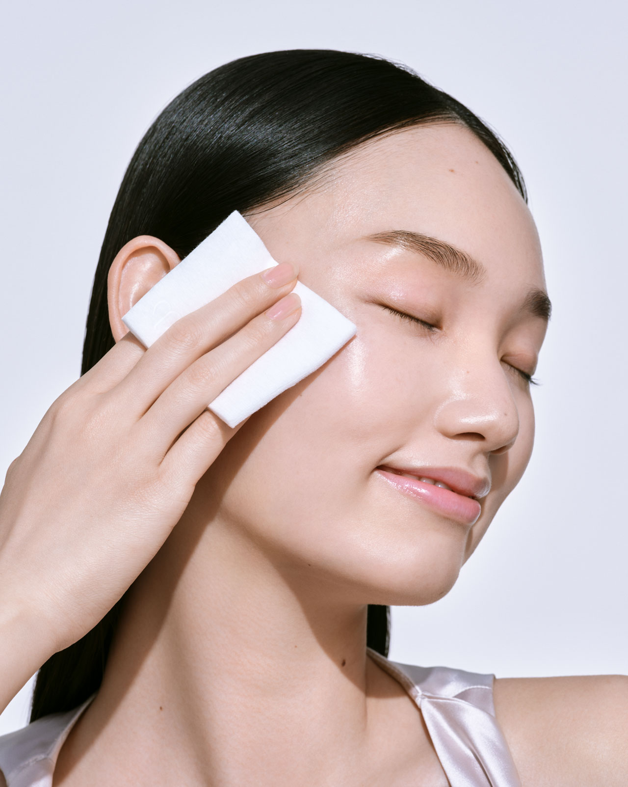 Photo of a woman gently gliding a white rectangular cotton pad over her face.