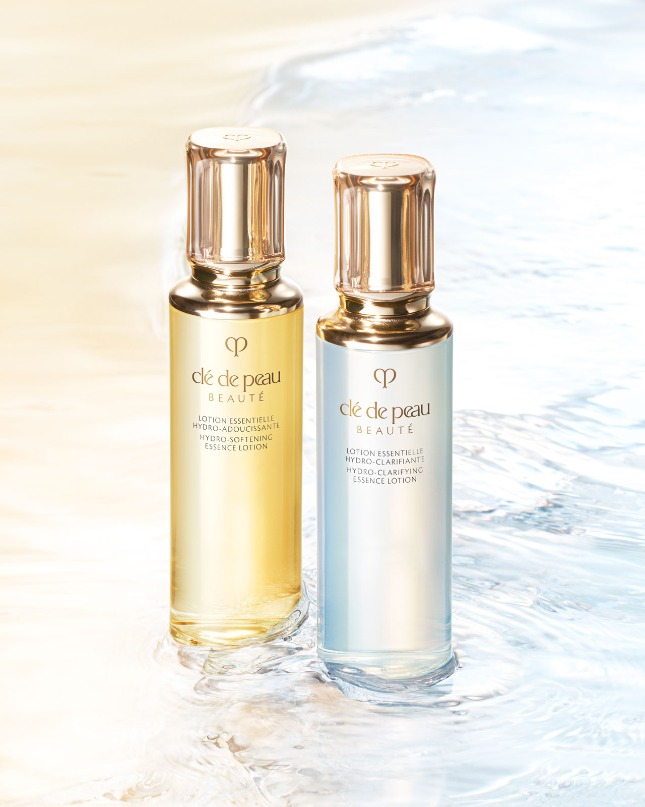 Photo of two essence lotions. On the left is HYDRO-SOFTENING ESSENCE LOTION in a slightly yellow-tinted bottle, and on the right is HYDRO-CLARIFYING ESSENCE LOTION in a clear bottle. Both bottles feature a translucent, rounded gold cap and a gold brand logo.