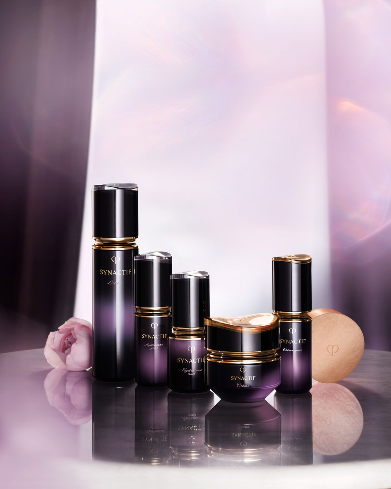 Photo of the Clé de Peau Beauté Synactif skincare lineup, arranged from left to right as: Lotion, Daytime Moisturizer, Nighttime Moisturizer, Cream, Eye Cream, and Soap — all in gradient black-to-purple packaging with gold accents.