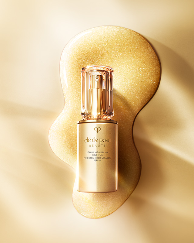 Photo of the product bottle of Clé de Peau Beauté's Precious Gold Vitality Serum, elegantly placed on a rich, shimmering swatch of the serum's 24K Gold-infused texture, conveying its luxurious quality.