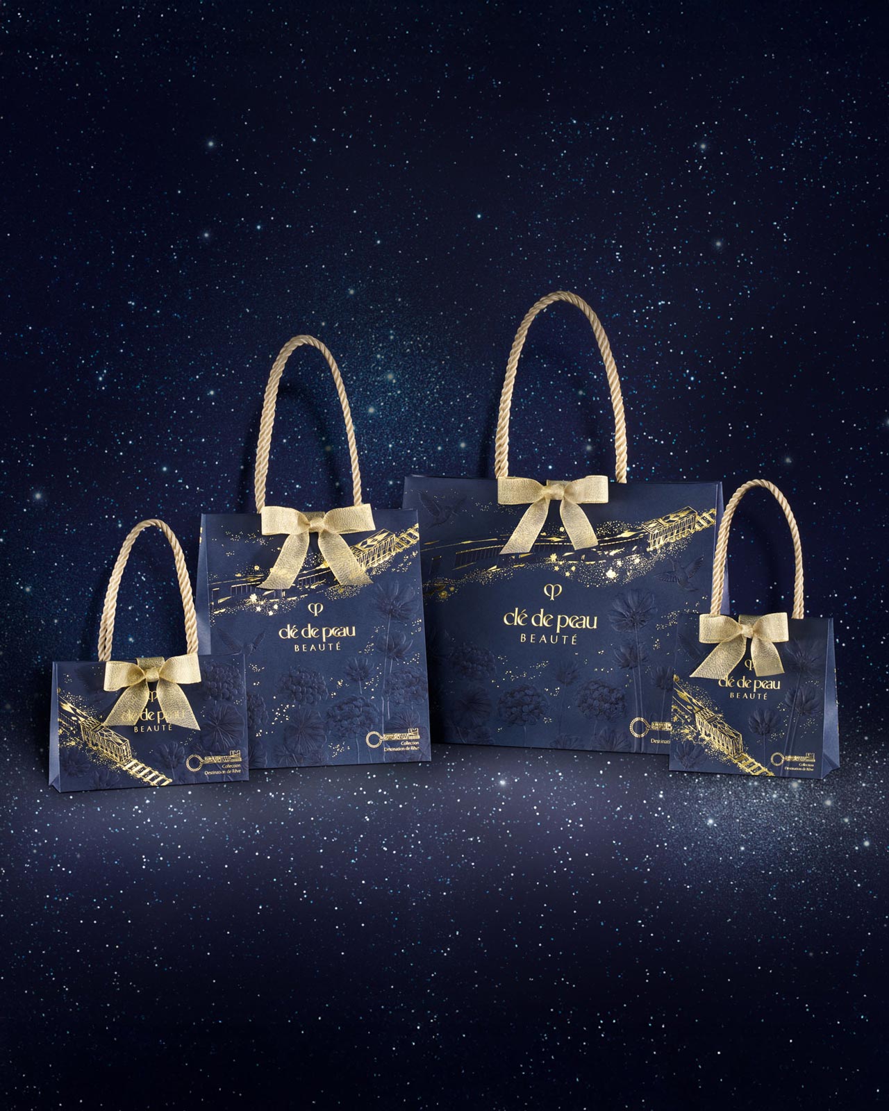 Photo of four limited-edition Clé de Peau Beauté gift bags of varying sizes, lined up to showcase the special holiday collection packaging. The bags feature a deep blue design with elegant gold ribbons.