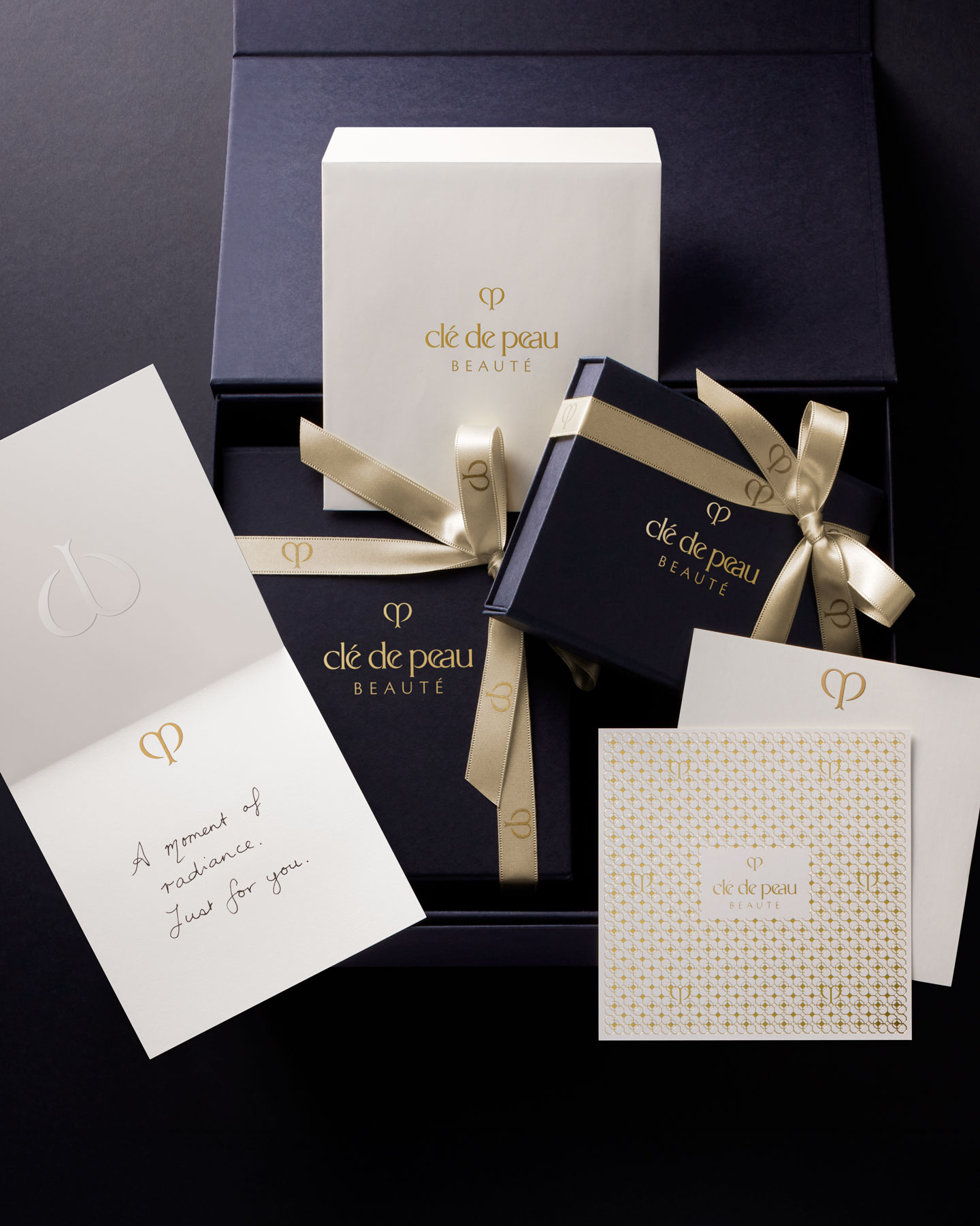 Photo of a sophisticated dark blue Clé de Peau Beauté gift box and its accompanying message card, both imprinted with the brand's logo, with the box adorned with a gold ribbon.