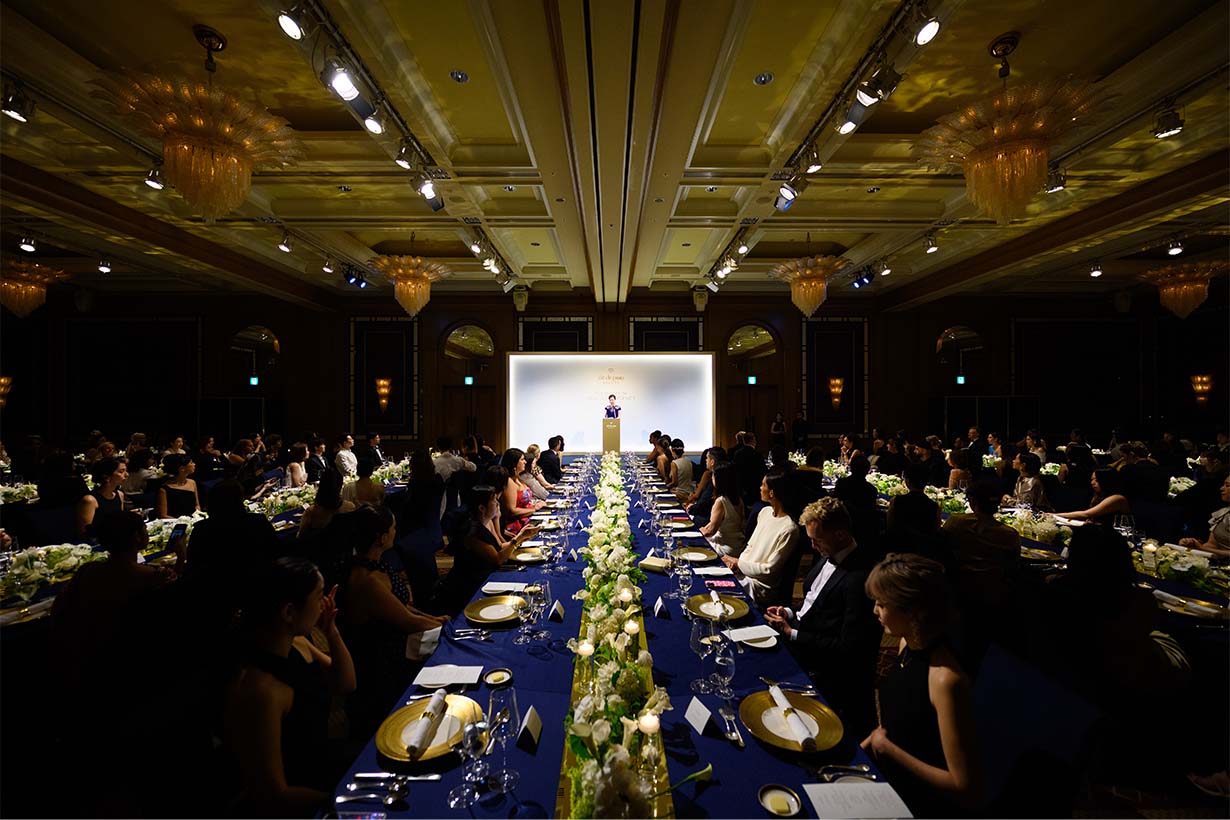 Photo of numerous guests seated at tables in an event hall, while a speaker presents on stage.
