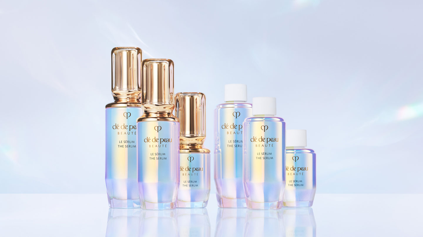 Photo of multiple bottles of Clé de Peau Beauté THE SÉRUM in various sizes, featuring iridescent packaging with gold or white caps.