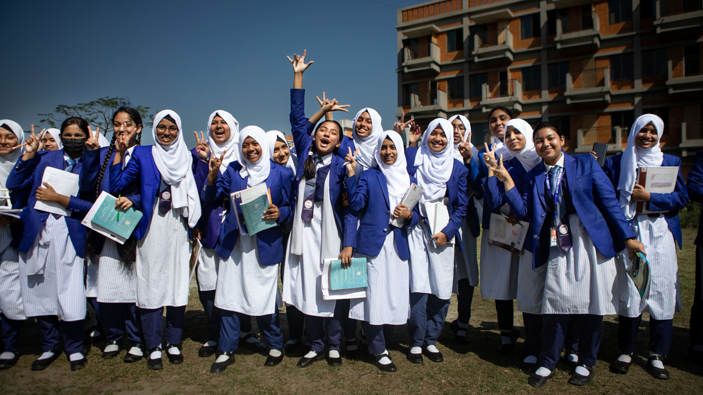 Photo of Bangladeshi schoolgirls in uniform smiling and holding textbooks outside their school building.